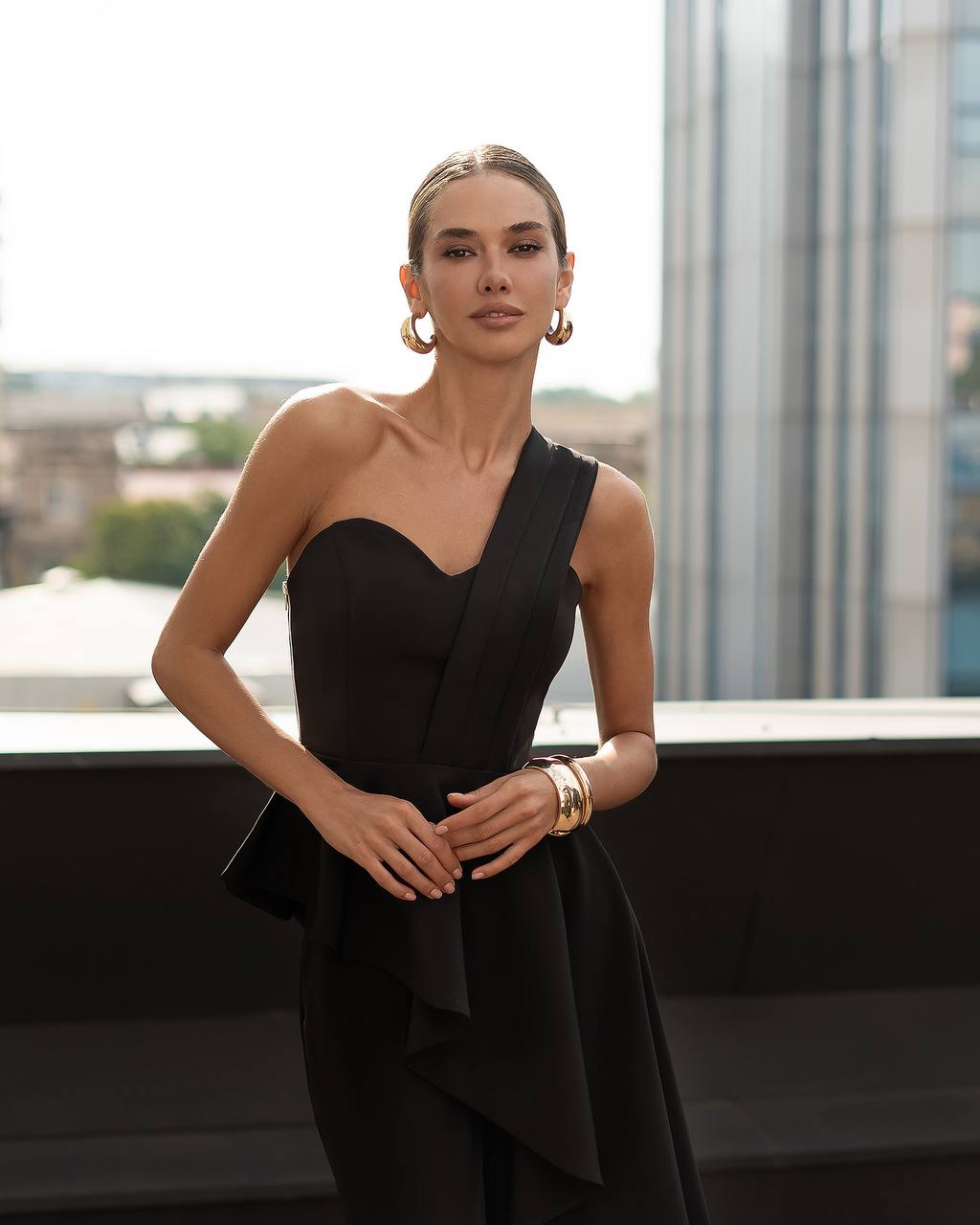 Woman in a black one-shoulder dress standing in front of a window with a cityscape view.