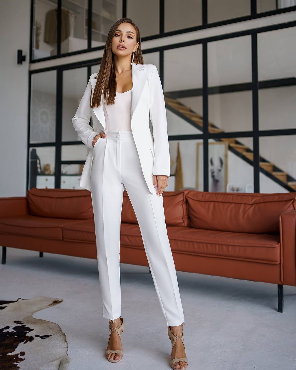 Woman in a white suit standing in a modern interior setting with a brown sofa and geometric wall design.