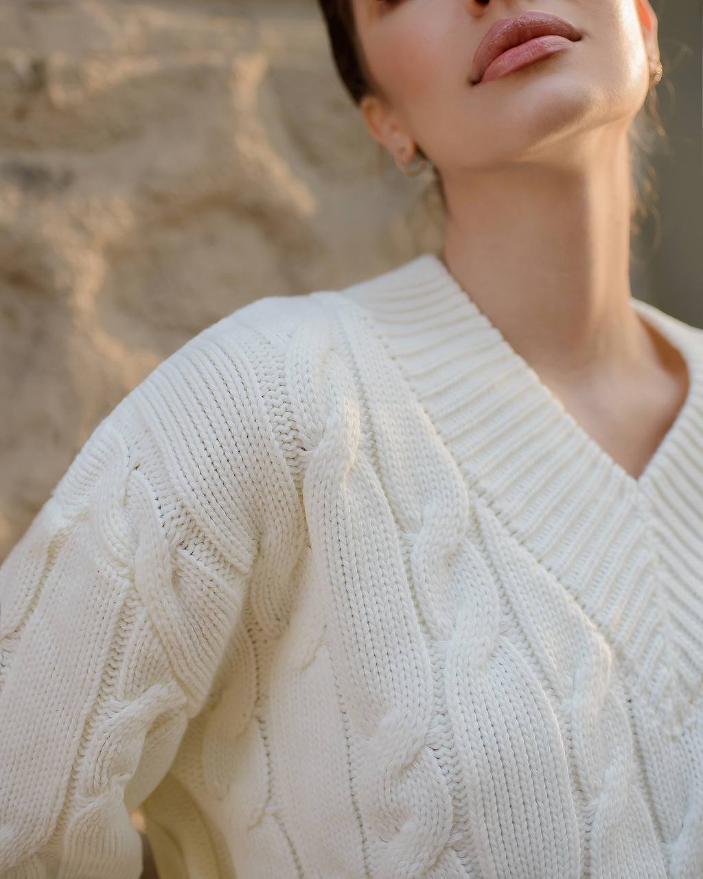 Close-up of a person wearing a cream-colored cable knit sweater against a blurred natural background.