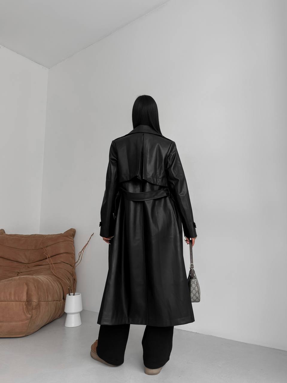Person wearing a black leather trench coat in a minimalistic room with a brown bean bag chair.