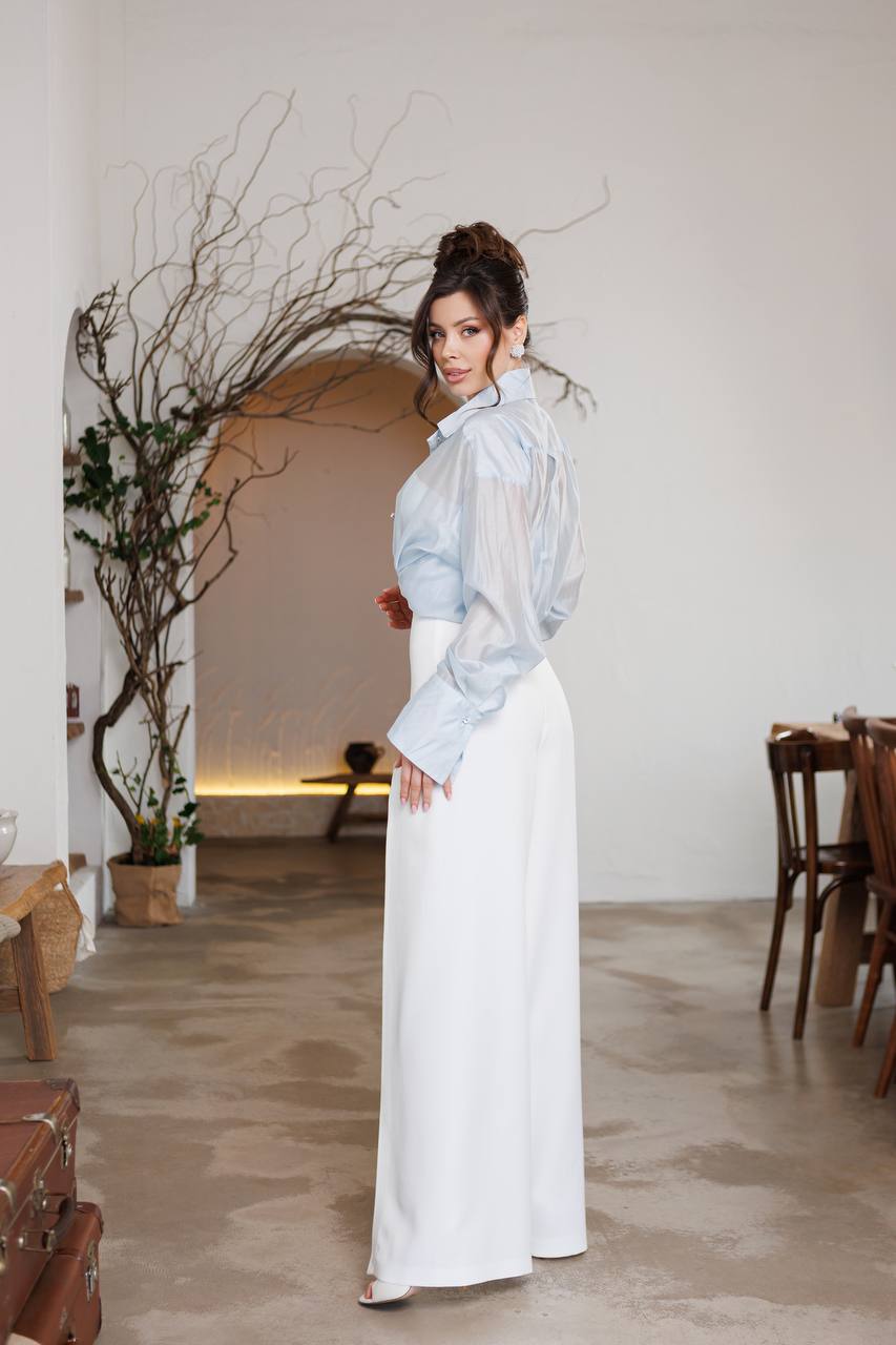 Woman in a light blue blouse and white pants standing in a room with a neutral color palette.