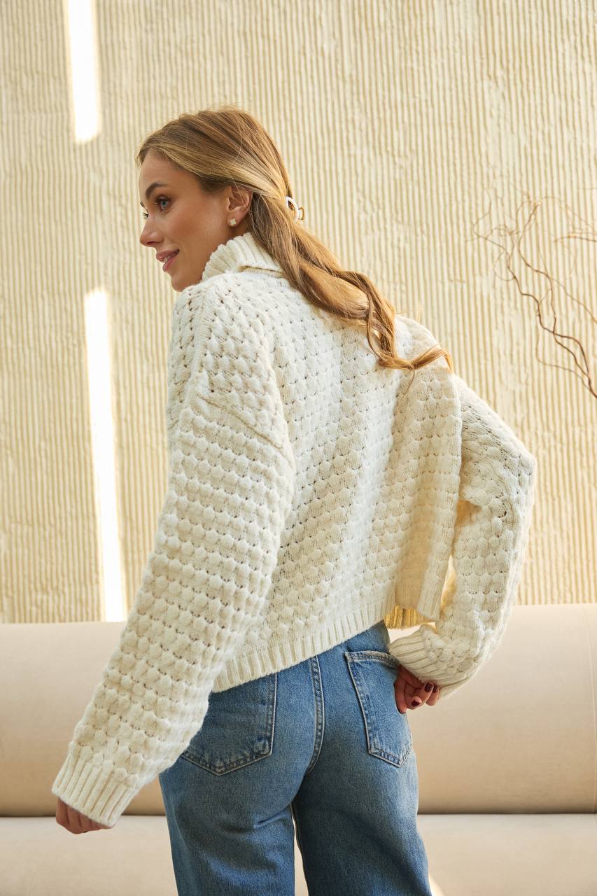 Woman wearing a white textured sweater and blue jeans indoors.