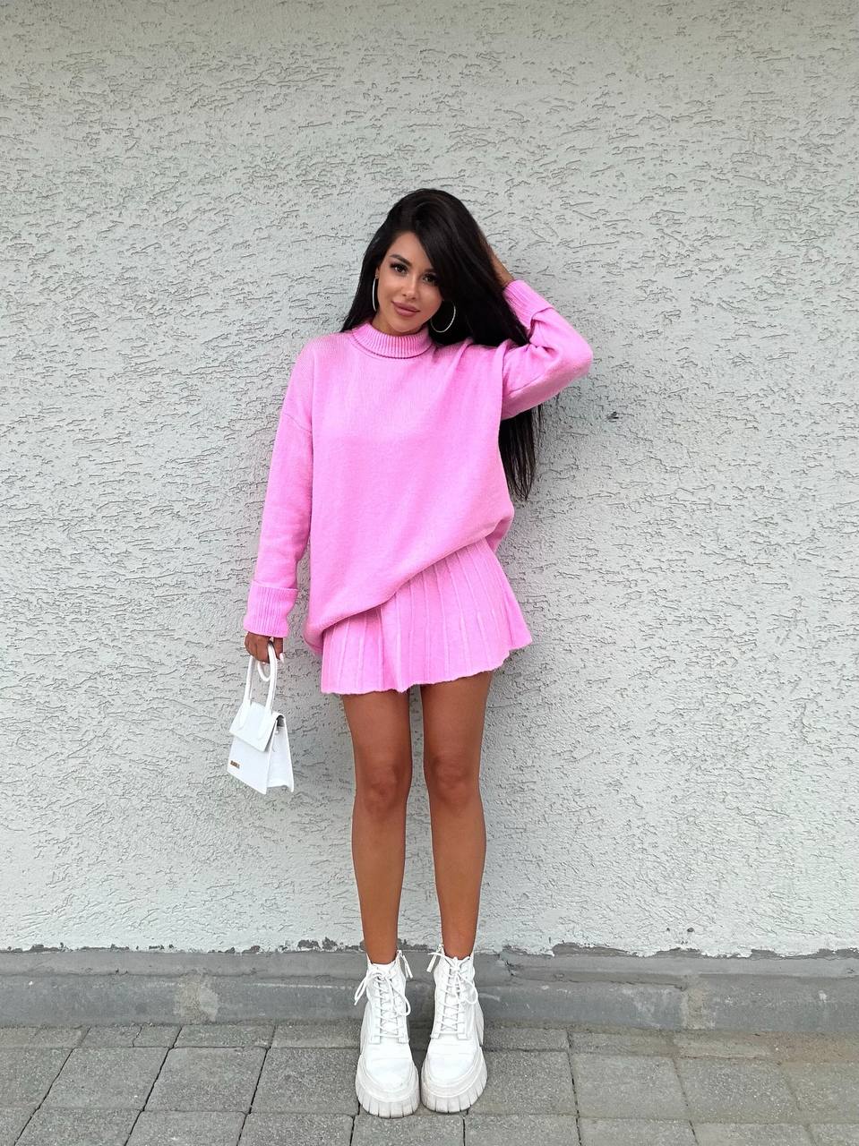 Woman wearing a pink outfit with a sweater and skirt against a gray wall.