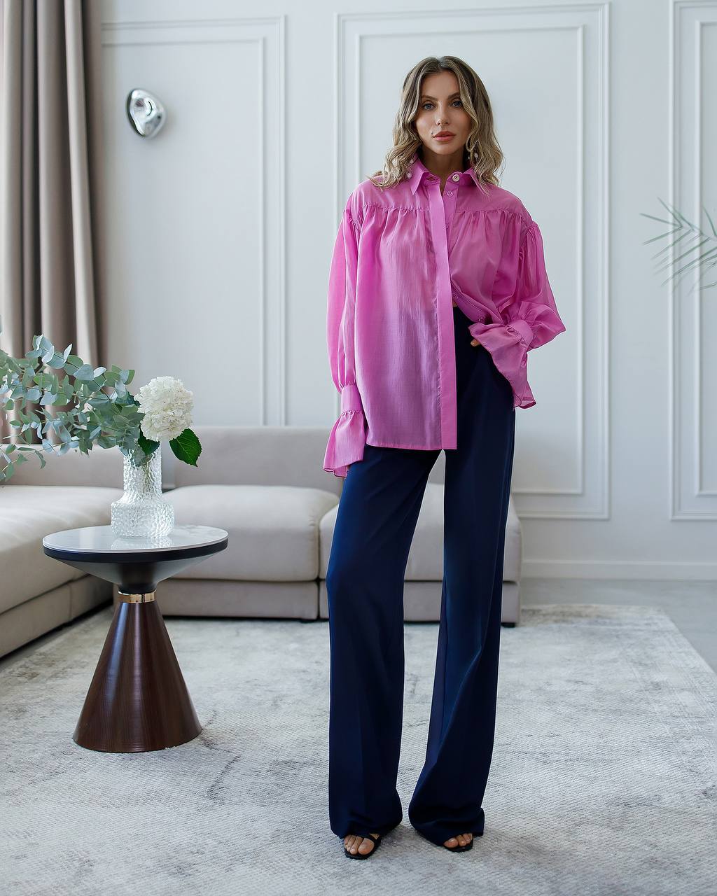 Woman wearing a pink blouse and dark pants in a living room setting.