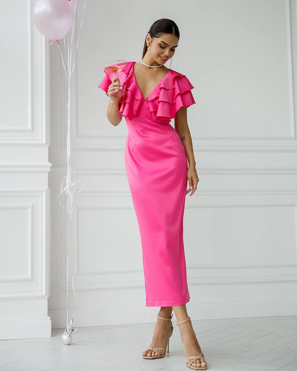 Woman in a bright pink dress holding a glass against a white wall with balloons.