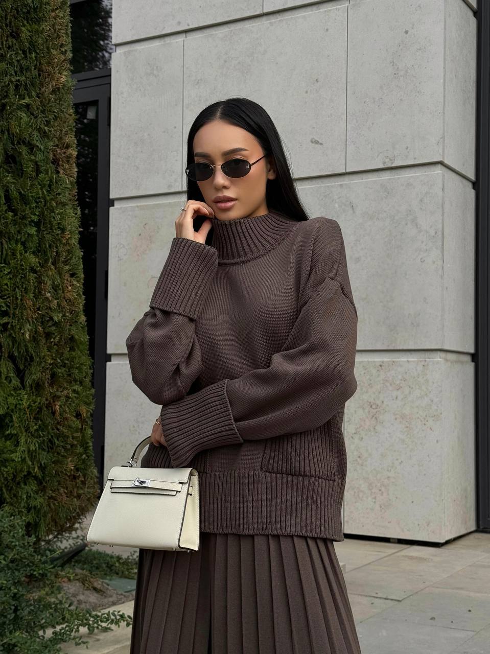 Woman wearing a brown sweater and skirt with sunglasses, standing against a light-colored wall.