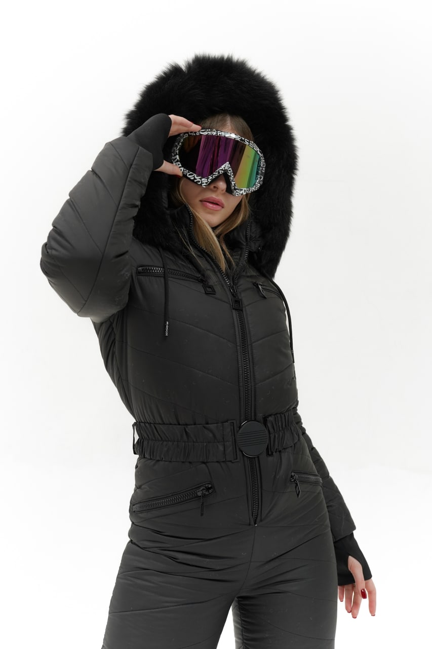 Person wearing a black snowsuit with a fur-lined hood, holding colorful goggles.