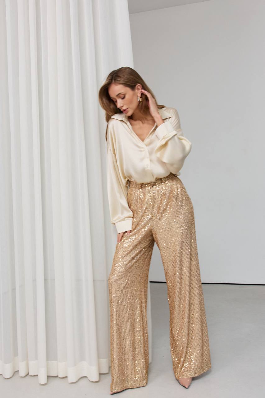 Woman wearing a cream blouse and gold sequin pants against a white curtain.