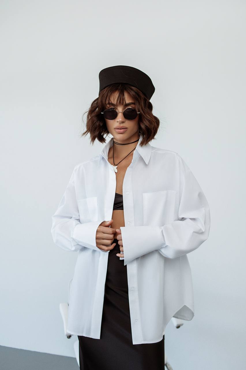 Person wearing a white shirt over a black outfit with sunglasses and a cap on a plain background