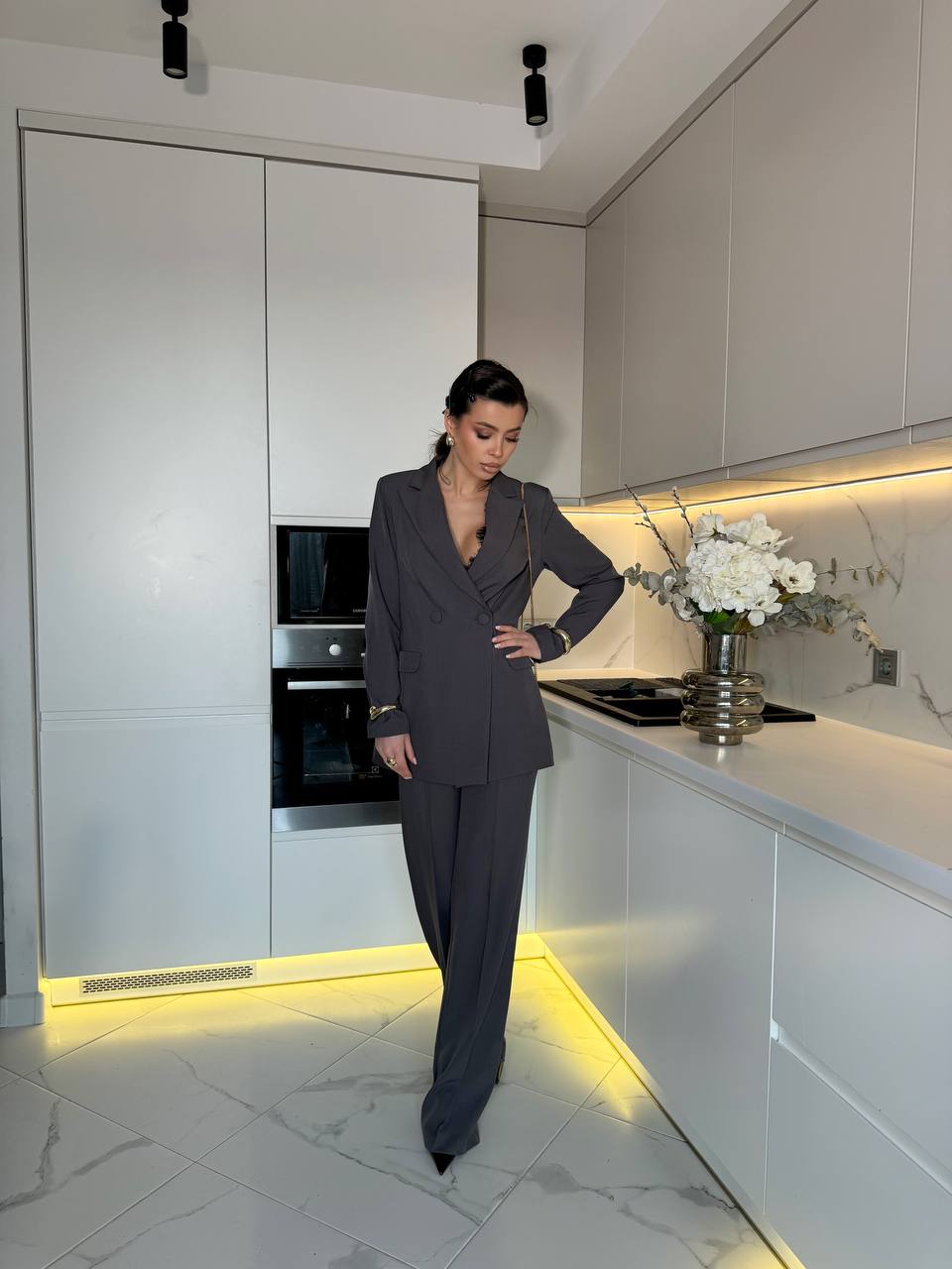 Woman in a gray suit standing in a modern kitchen with white cabinets and marble floor.