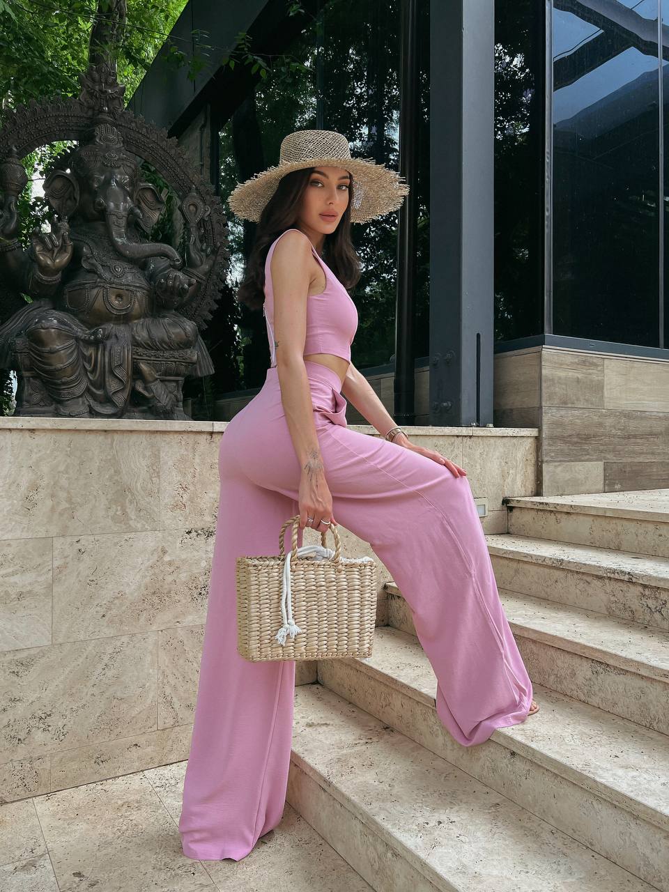 Woman in a pink outfit sitting on steps holding a straw bag, with a statue and building in the background.