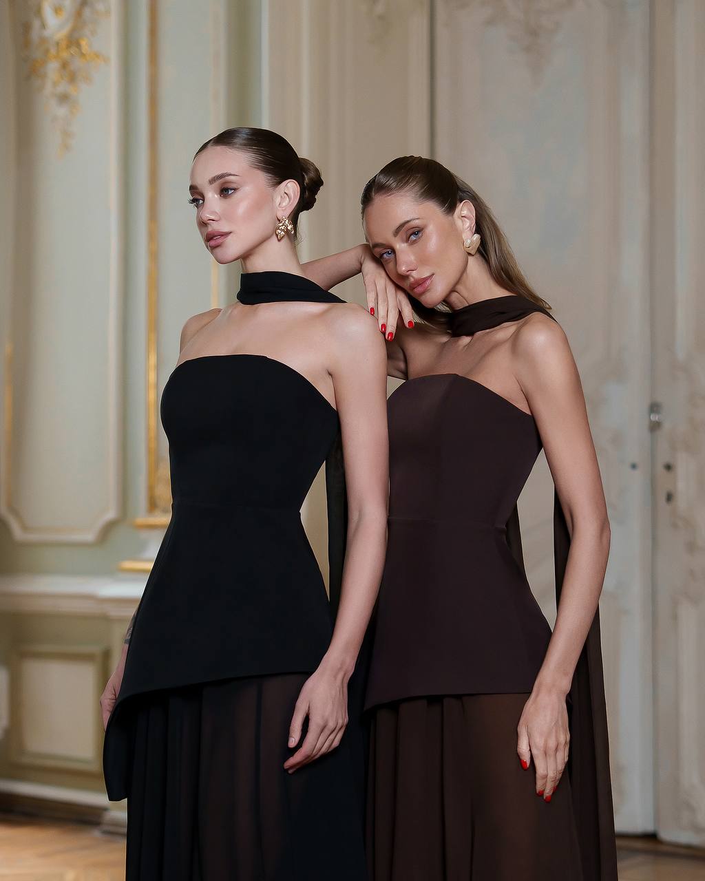 Two women wearing black halter neck dresses in an elegant interior setting.