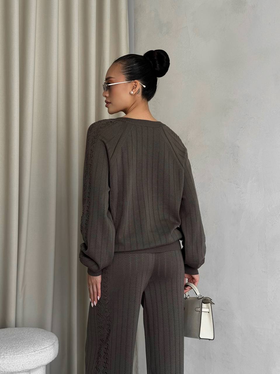 Woman in a brown outfit standing against a neutral wall with a curtain.