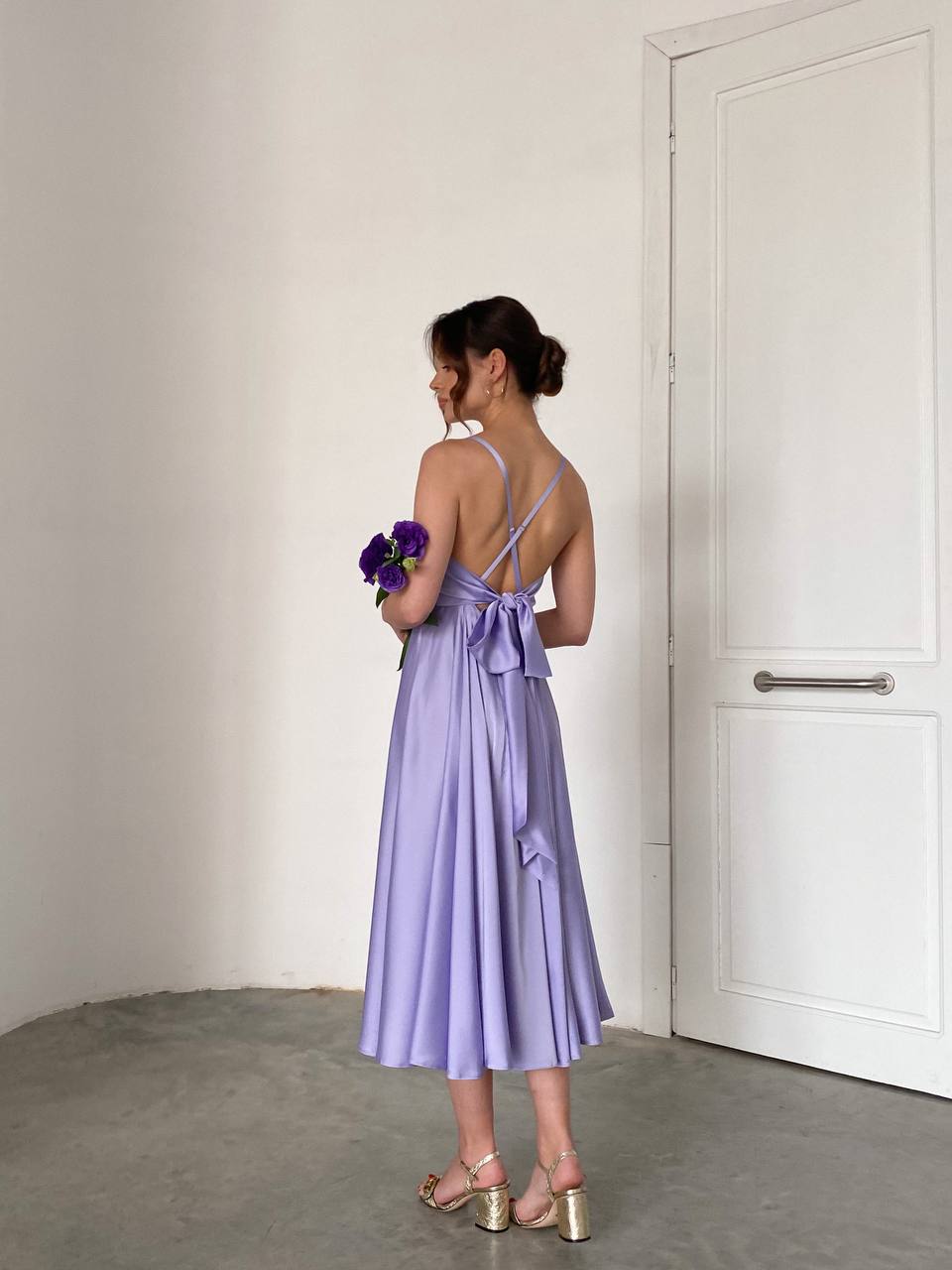 Woman in a lavender dress holding flowers against a plain wall.
