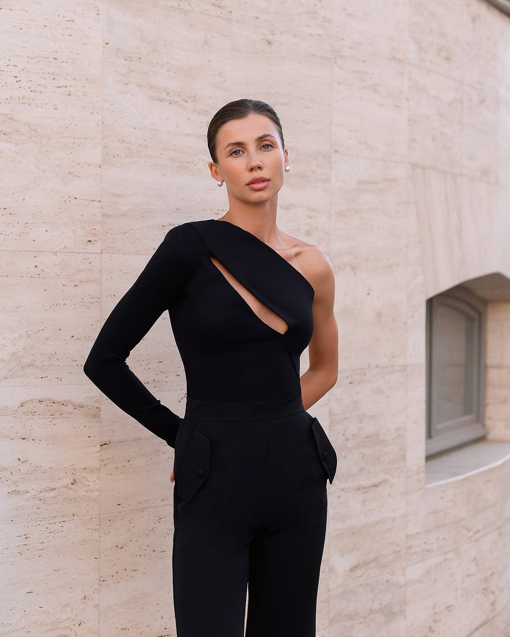 Woman wearing a black one-shoulder outfit against a beige wall.