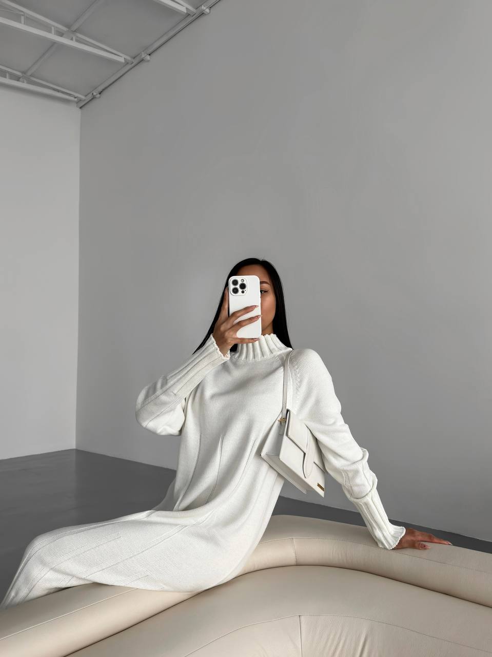 Person wearing a white outfit sitting on a light-colored surface against a plain gray wall.