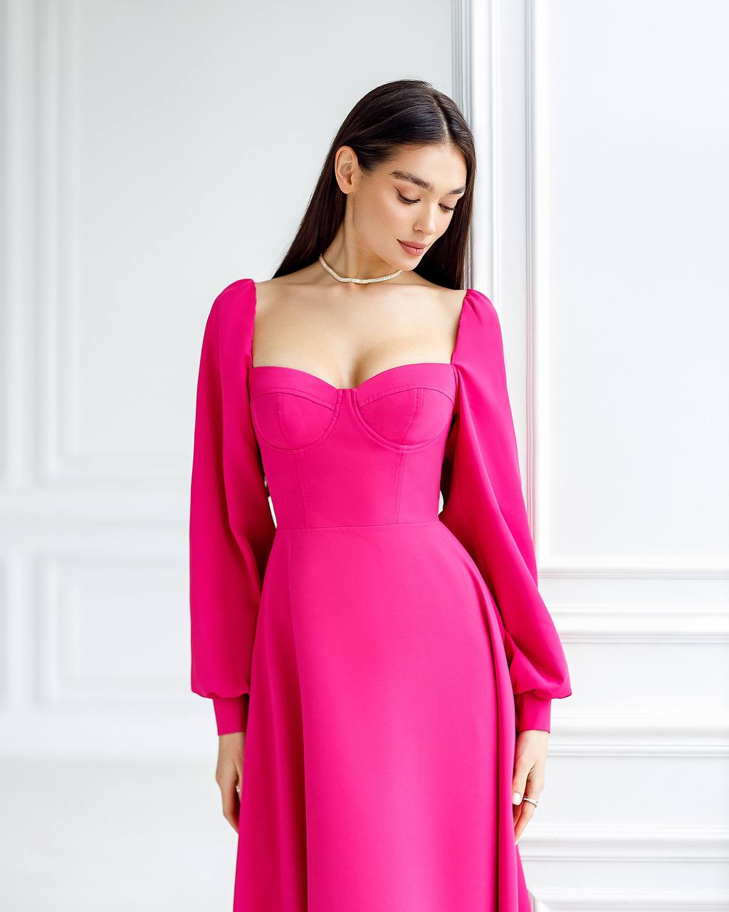 Woman wearing a bright pink dress with long sleeves against a white wall.