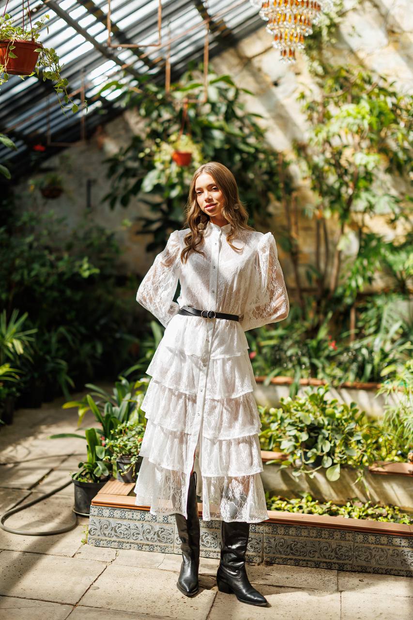 Woman in a white dress standing in a sunlit greenhouse filled with plants.