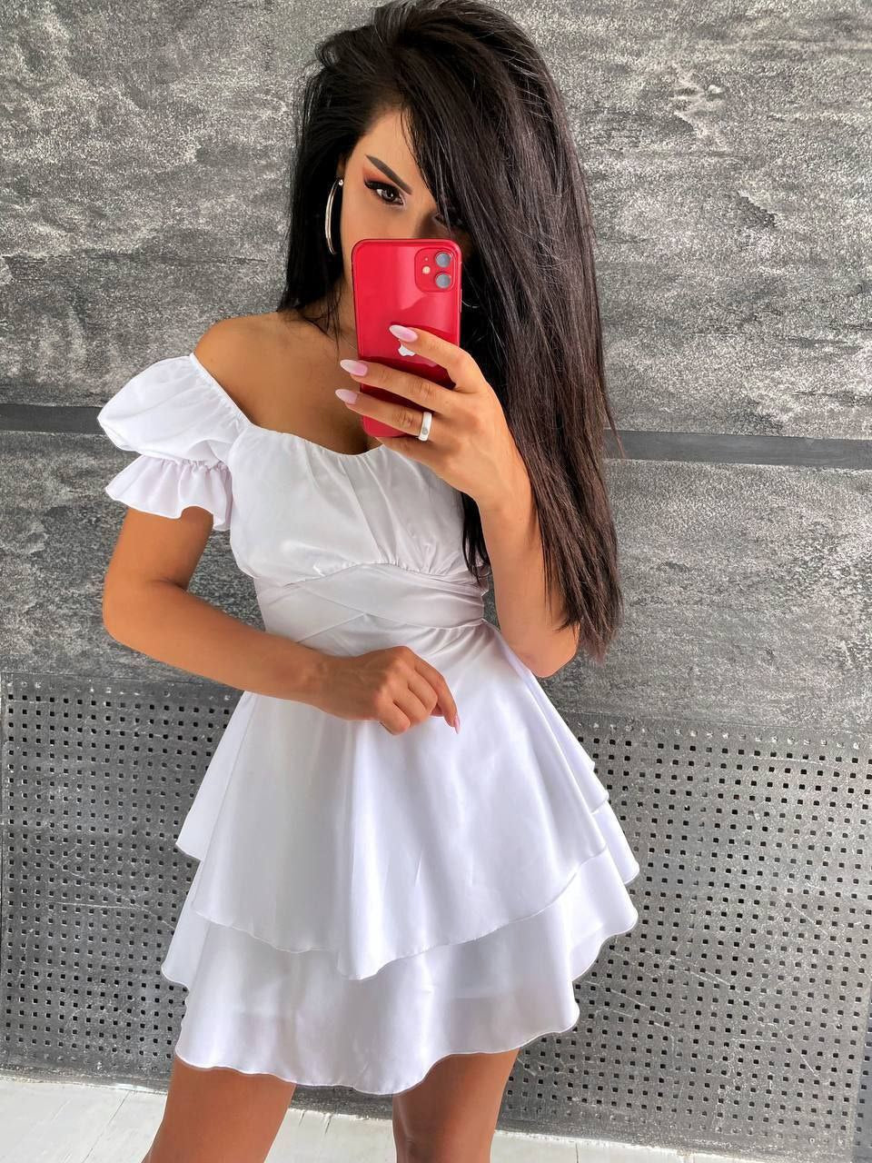 Woman taking a mirror selfie wearing a white dress against a gray wall.