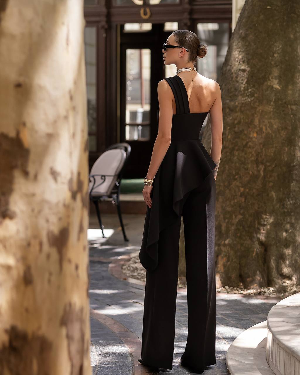 Woman in a black outfit standing in an outdoor setting with trees and chairs.