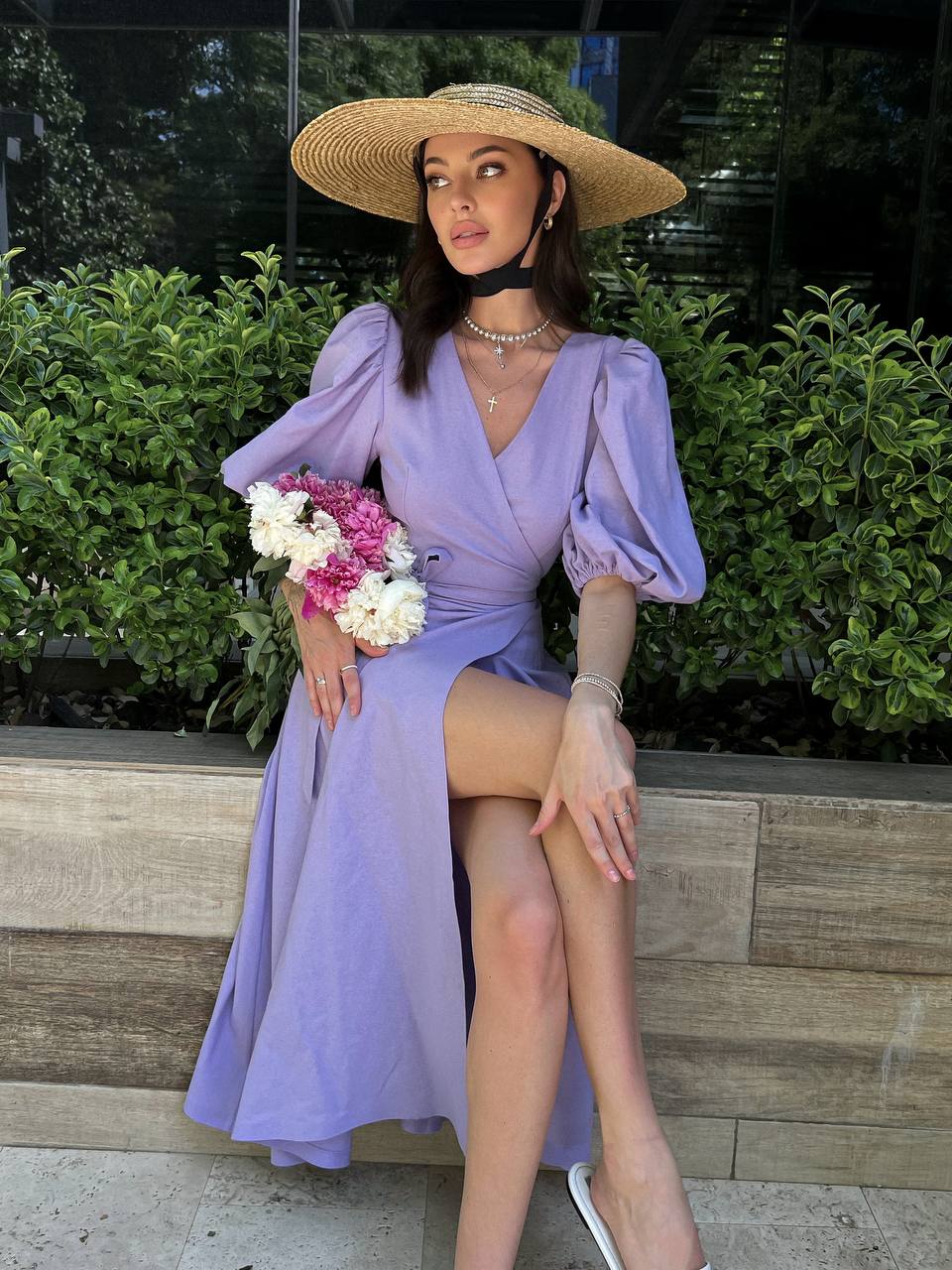Woman in a lavender dress and straw hat holding flowers, sitting on a stone ledge with greenery in the background.
