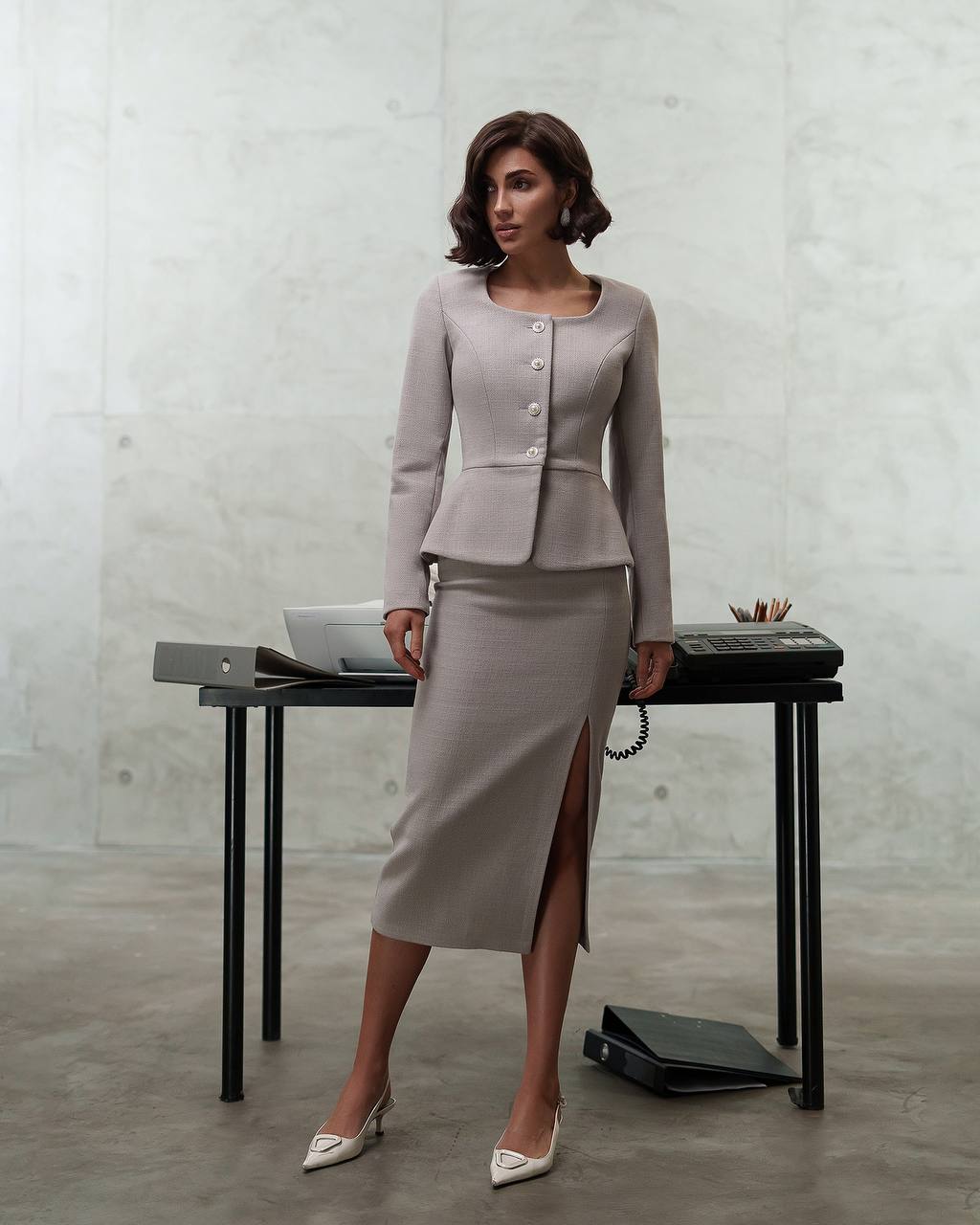Woman in a gray suit standing next to a desk with a concrete wall background