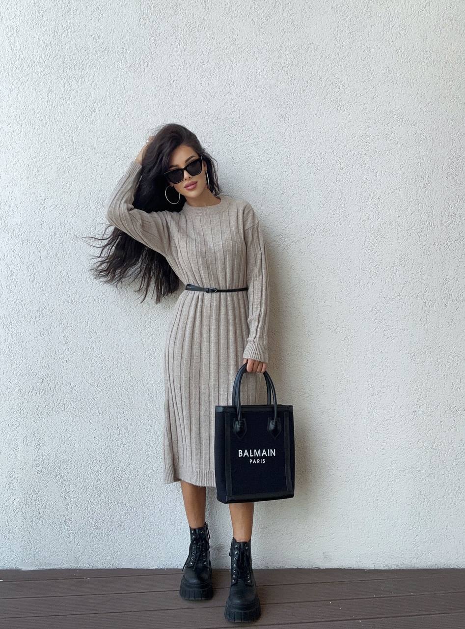 Woman in a beige dress holding a Balmain black handbag against a white wall.