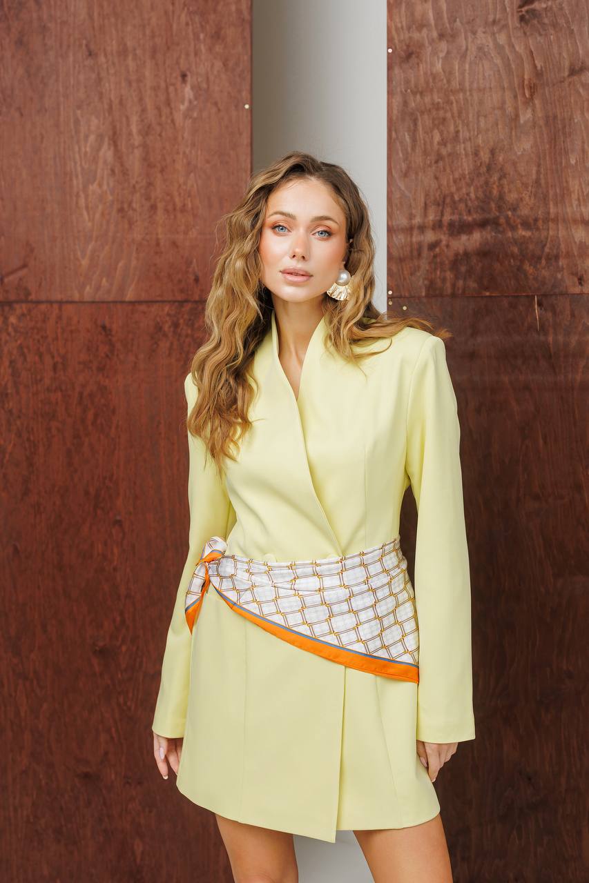 Woman in a light yellow outfit with a patterned sash against a brown wall.