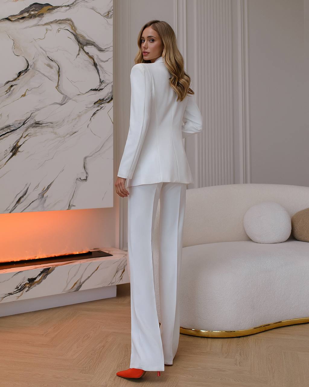 Woman in a white suit standing in a stylish room with marble wall and modern furniture.