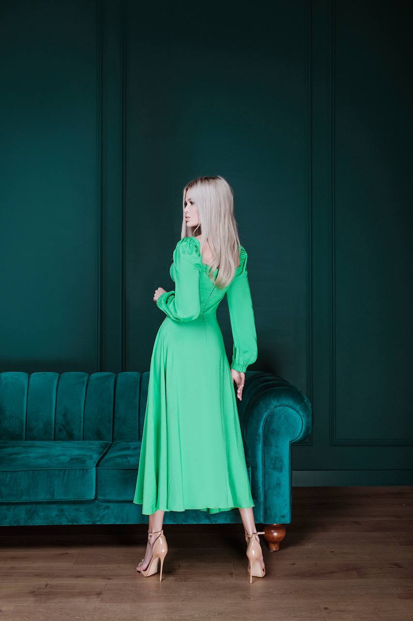 Woman in a green dress standing in a room with a teal sofa and dark walls.