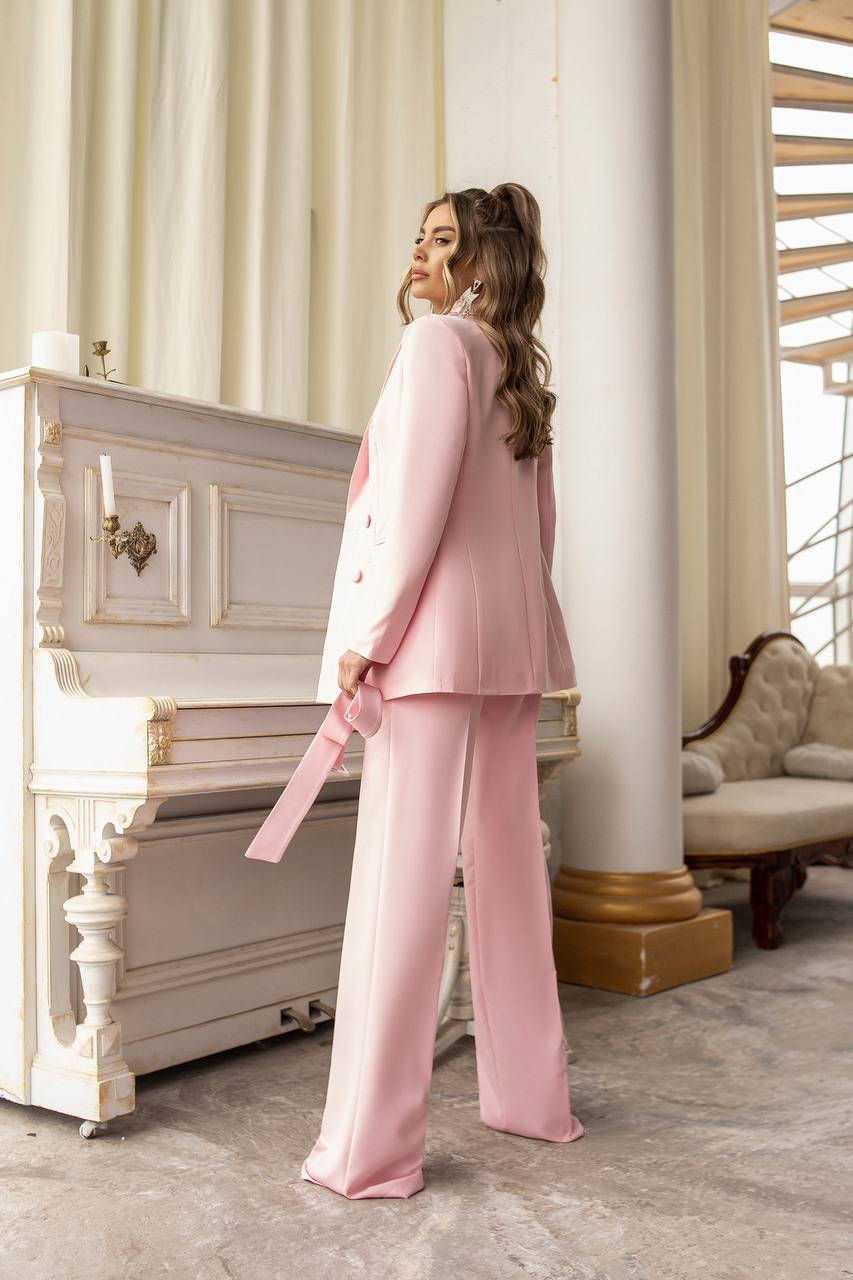 Woman in a pink suit standing in an elegant room with a piano and decorative columns.