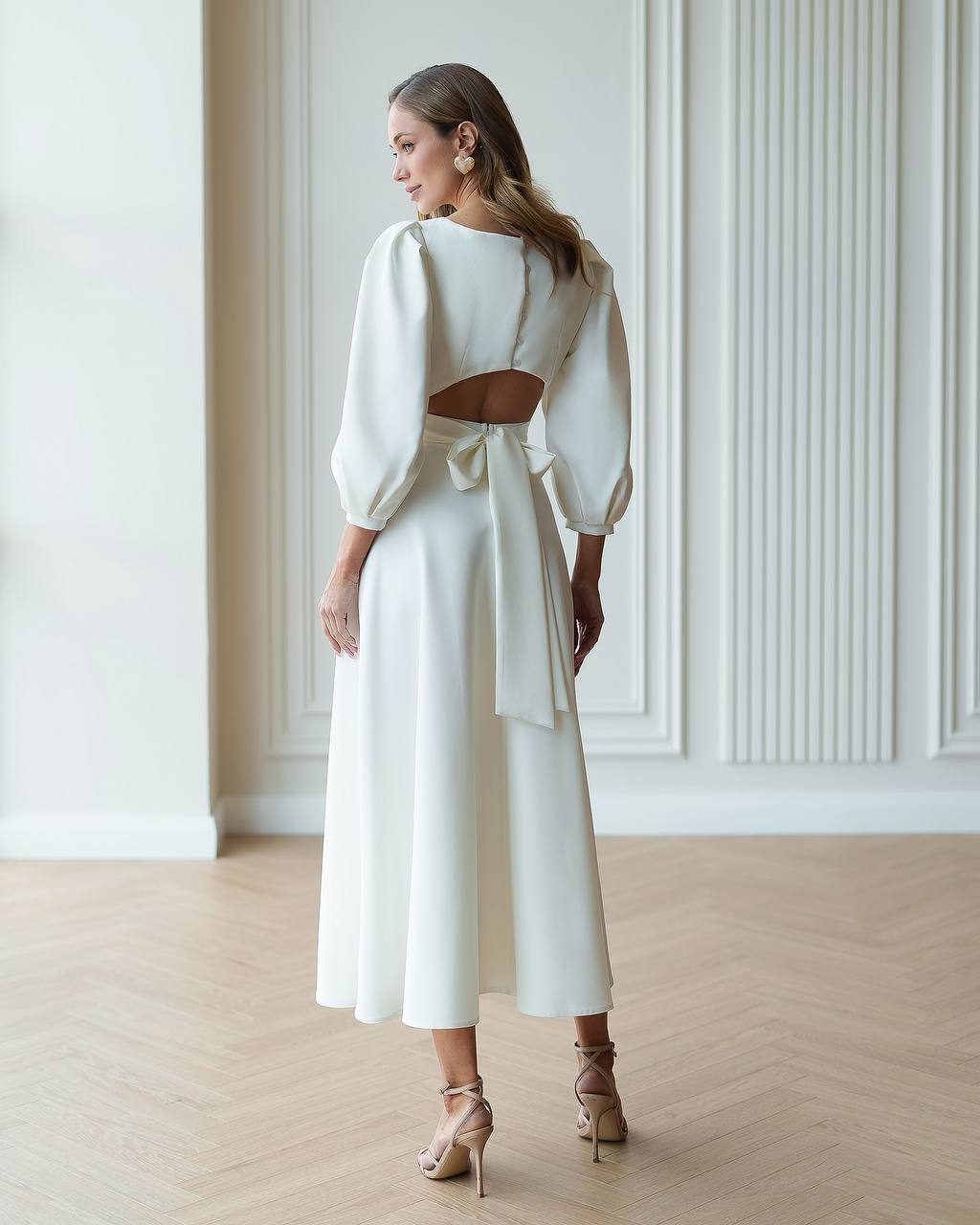 Woman wearing a white outfit with cut-out design in a room with wooden floor and white walls.