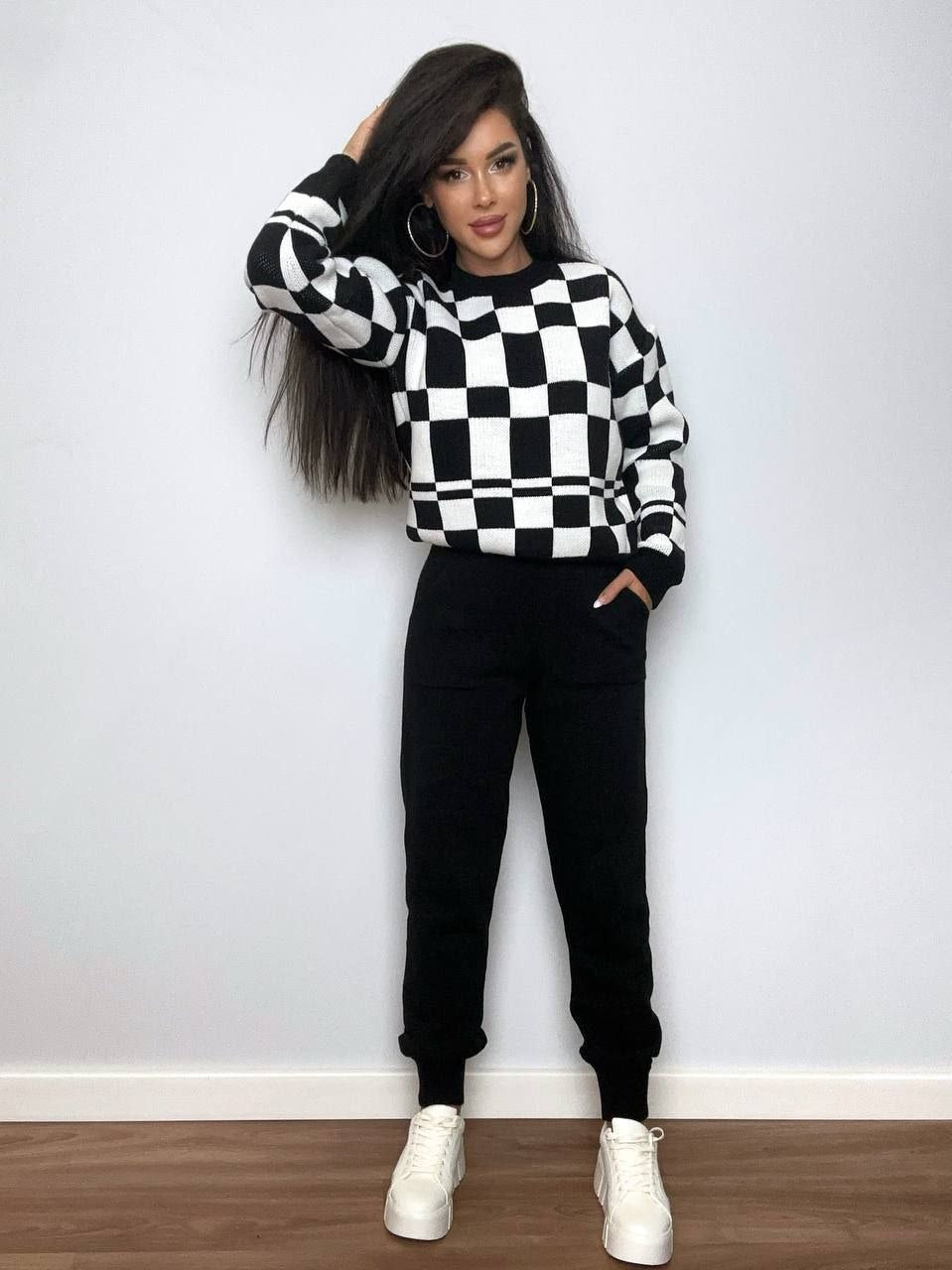 Woman wearing a black and white checkered sweater and black pants against a plain wall.