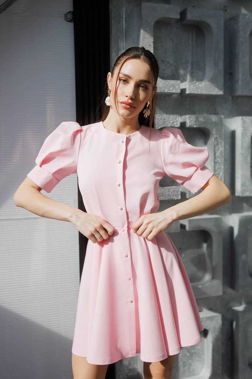 Woman wearing a pink dress with puffed sleeves in front of a textured wall.