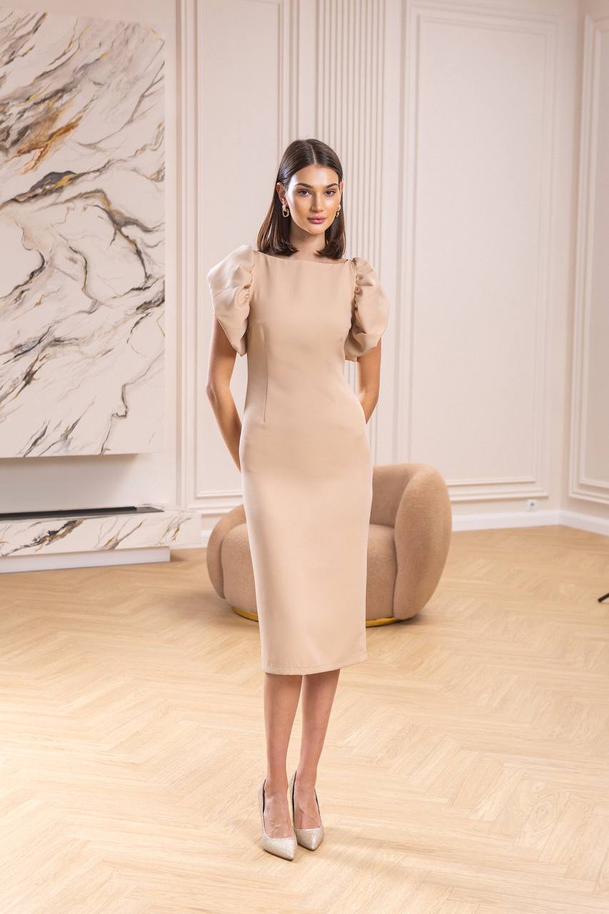 Woman in a beige dress standing in a room with marble wall and modern furniture.