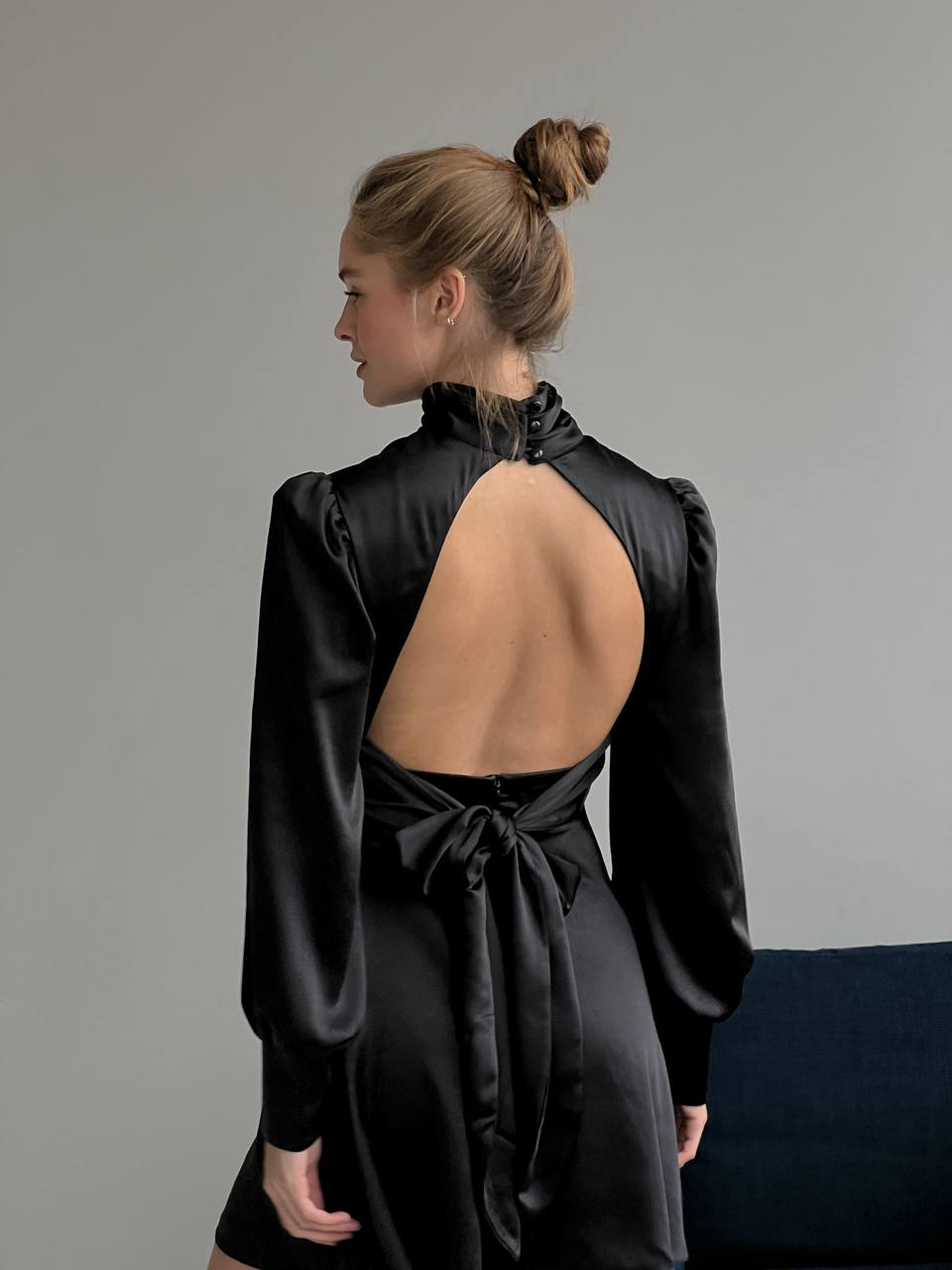 Woman wearing a black satin dress with an open back and bow detail, standing against a plain background.