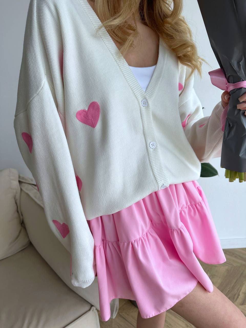 Person wearing a white cardigan with pink heart patterns and a pink skirt indoors.