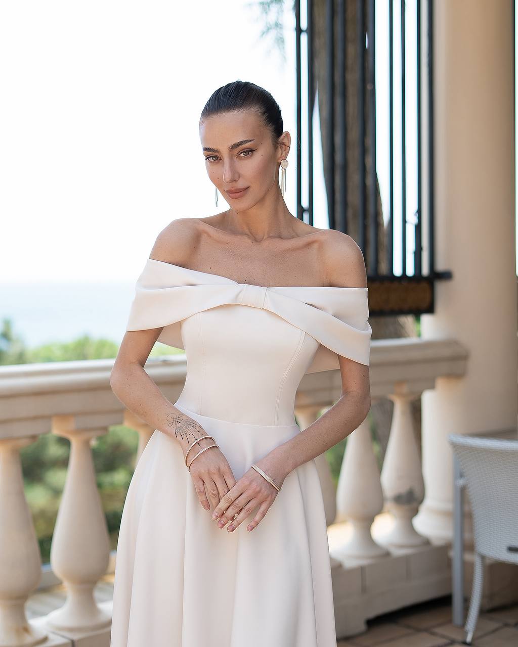 Woman in a white off-shoulder dress standing on a balcony.