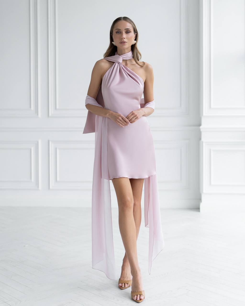 Woman wearing a light pink halter neck dress with sheer overlay against a white paneled wall.