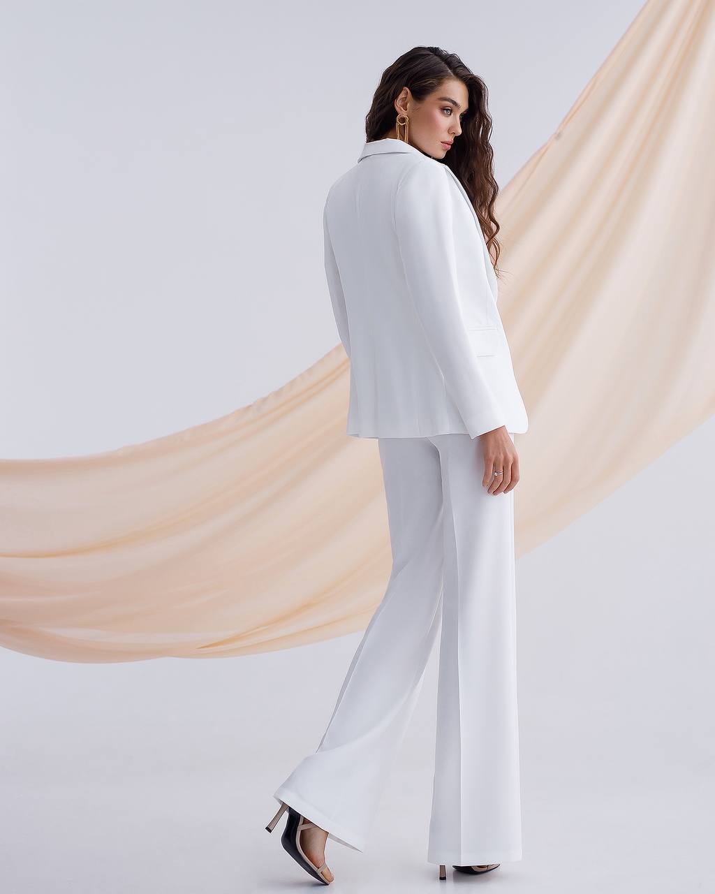 Woman wearing a white suit standing against a light background with draped fabric.