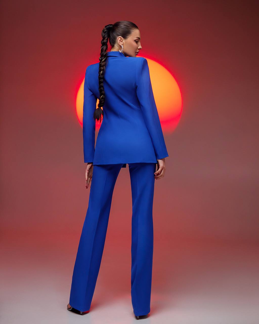 Woman in a blue suit standing with her back to the camera against a red and orange gradient background.