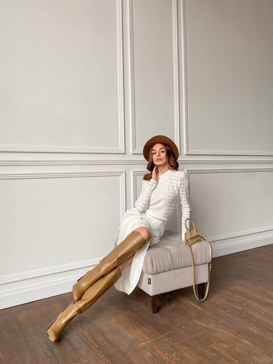 Woman sitting on a chair wearing a white outfit, brown boots, and a brown hat in a room with white paneled walls.