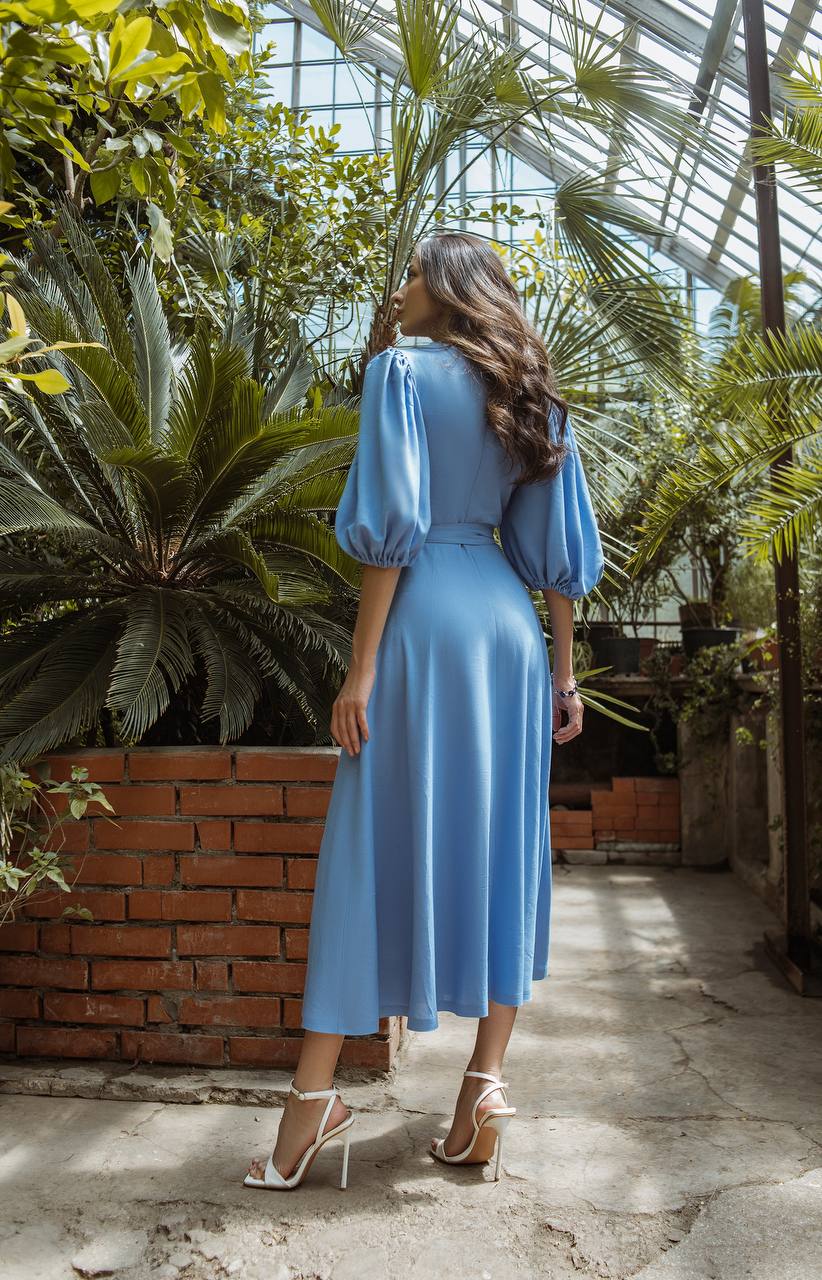 Woman in a blue dress standing in a greenhouse with plants and brick wall.