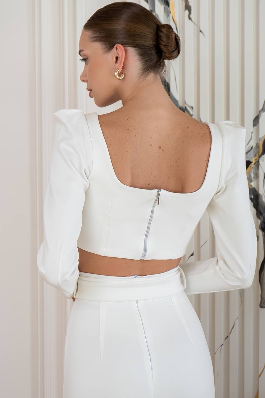 Woman wearing a white outfit with a backless top and high-waisted pants, standing against a neutral background.