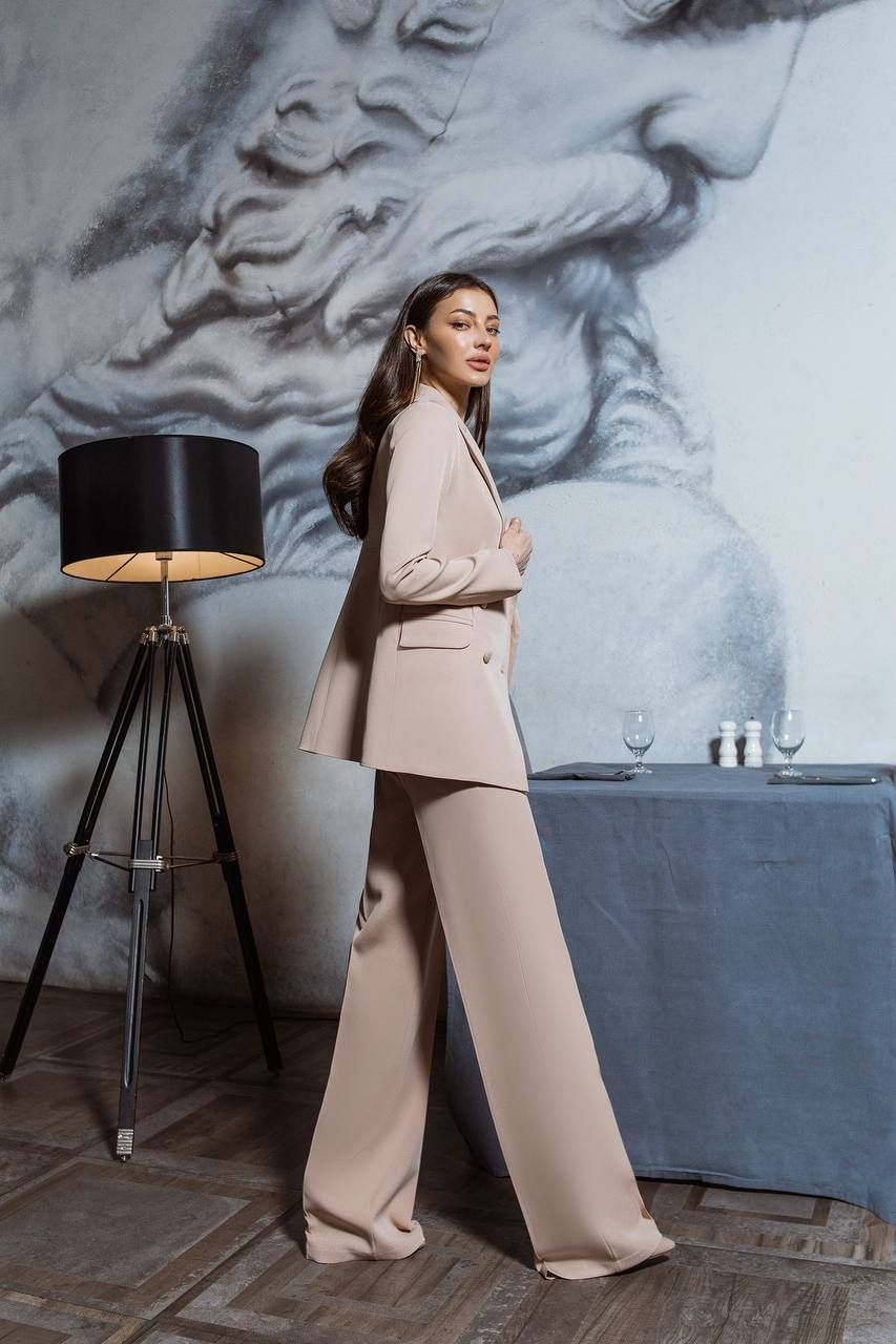 Woman in a beige suit standing in a room with a lamp and table in the background.
