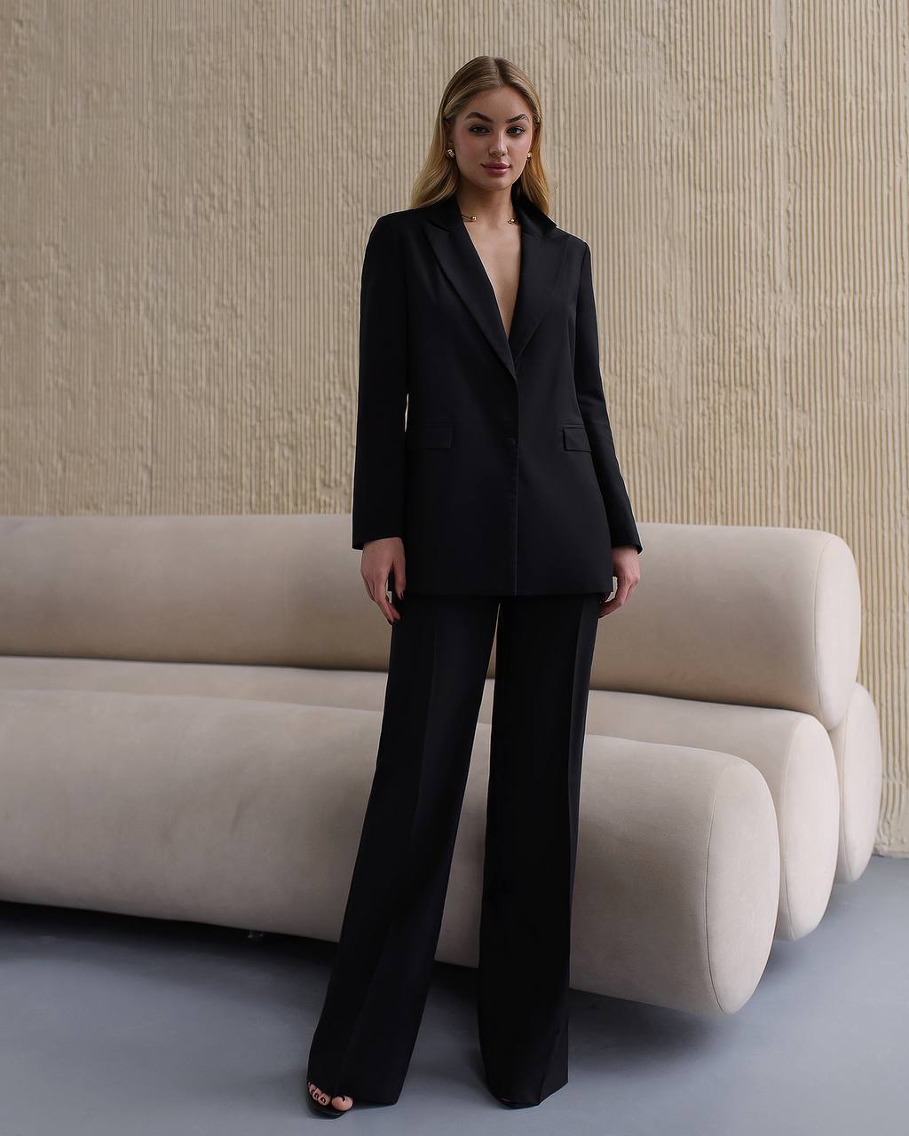 Woman in a black suit standing in a modern interior setting with a beige sofa.
