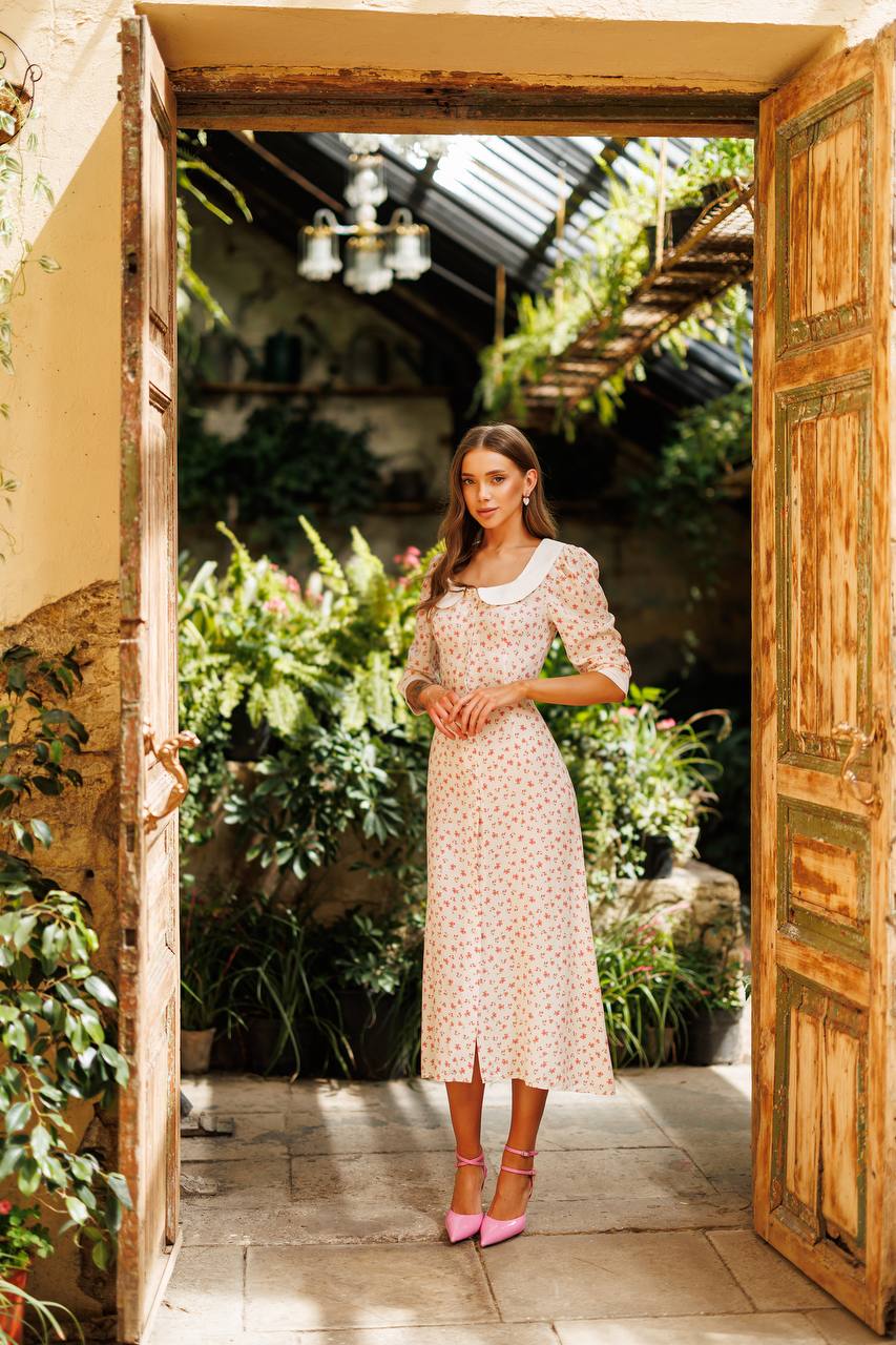 Woman in a floral dress standing in a sunlit garden with open wooden doors.