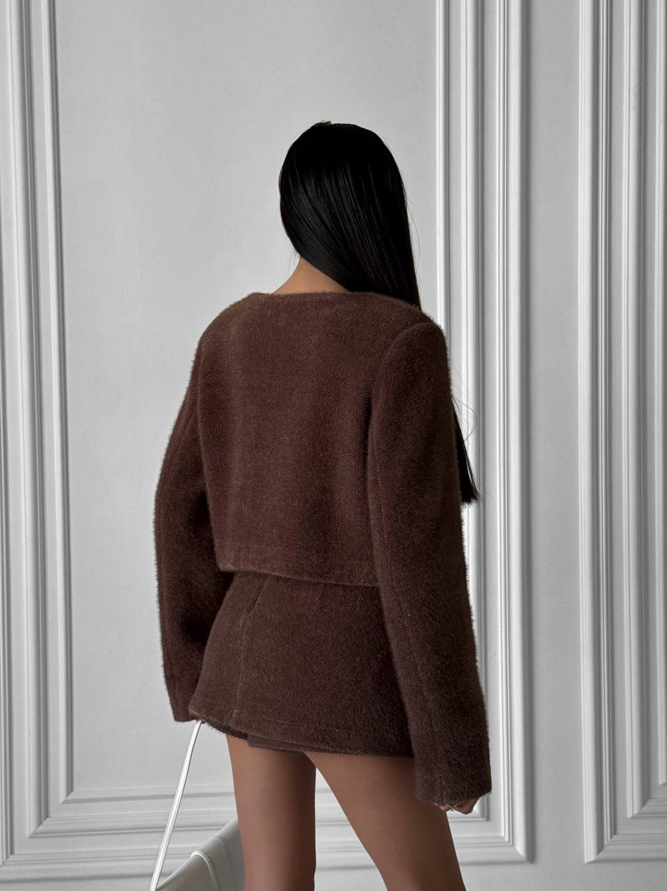 Person wearing a brown textured jacket standing against a gray paneled wall.