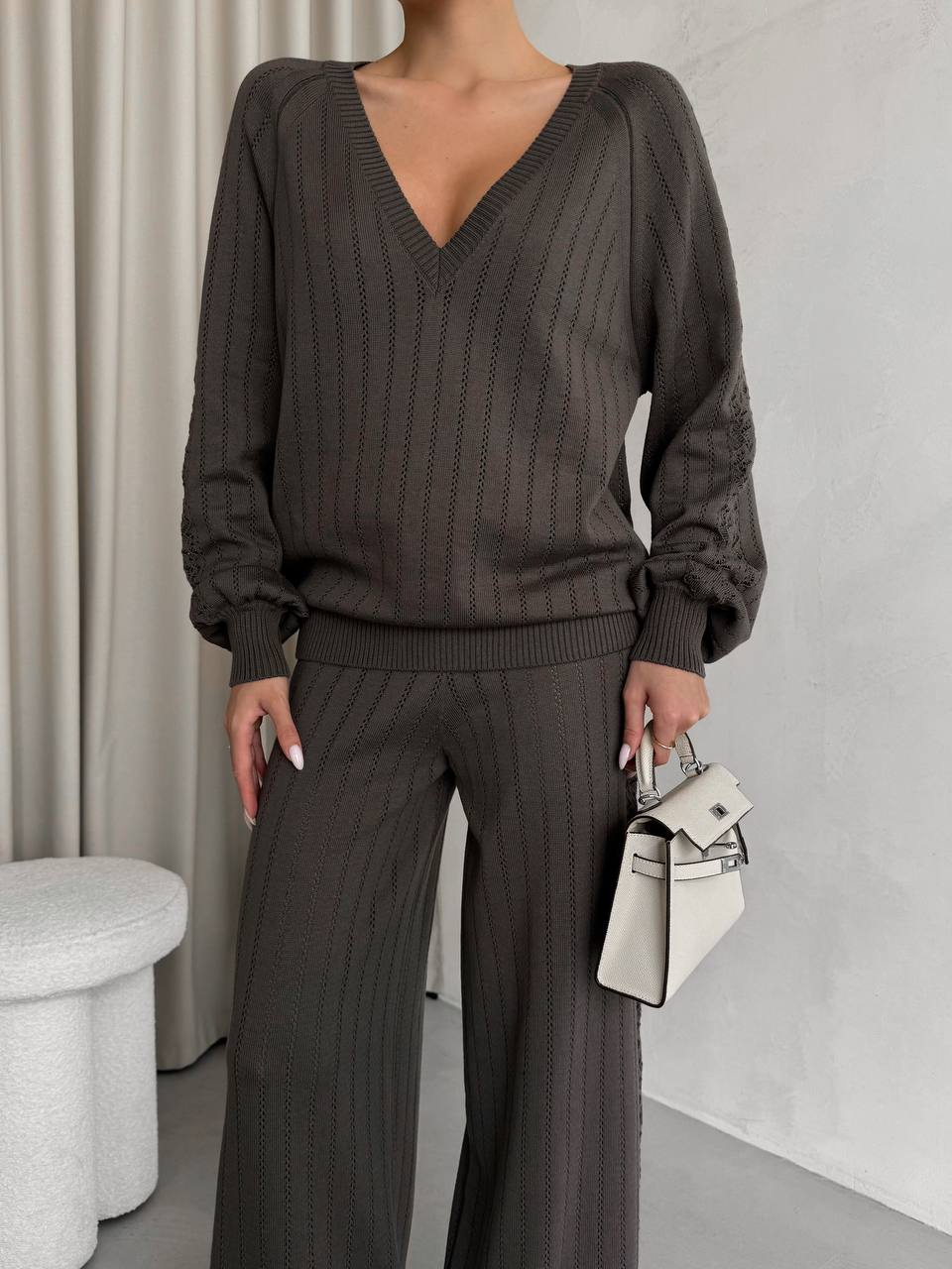 Person wearing a brown ribbed sweater and pants holding a white handbag indoors.