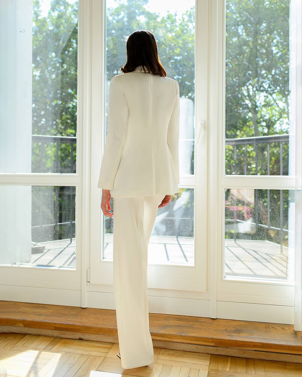 Person in a white suit standing in a room with large windows and a view of greenery outside.
